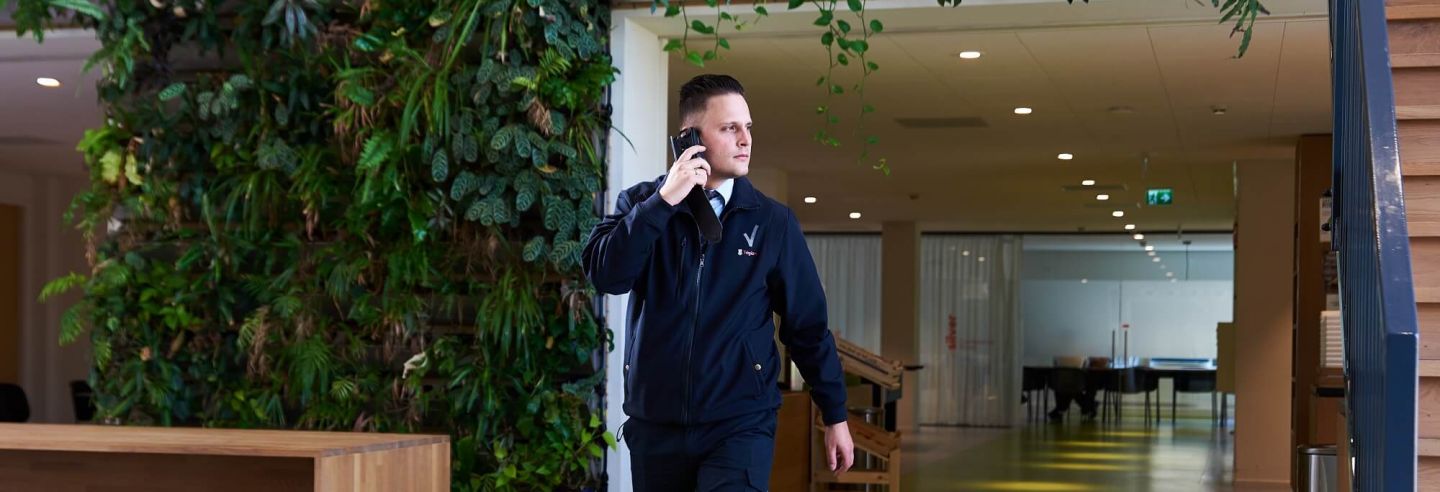Mannelijke beveiliger loopt in zijn uniform door gang met een groene omgeving en belt met de centrale over de ingekomen melding via een portofoon.