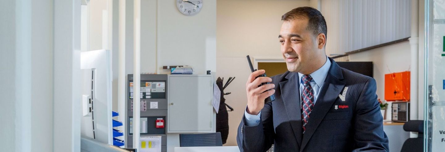 Mannelijke beveiliger in uniform praat via de portofoon met een collega beveiliger op locatie en kijkt daarnaast ook gelijk op zijn computerscherm achter zijn bureau.