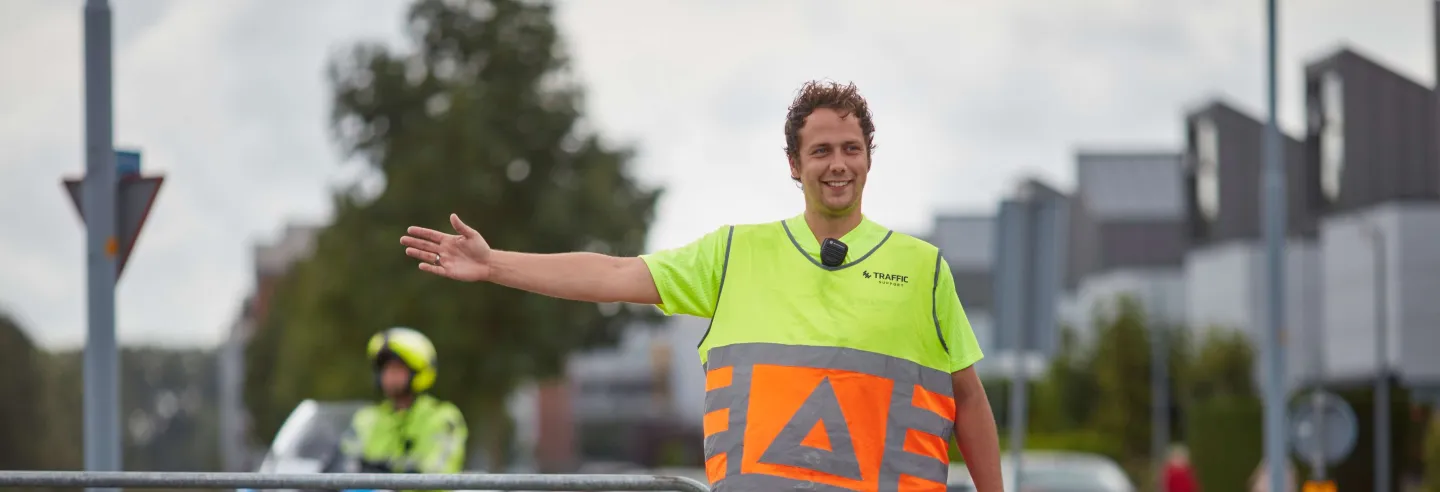 Verkeersregelaar in oranje en geel uniform strekt rechterarm uit en begeleidt het verkeer naar rechts.