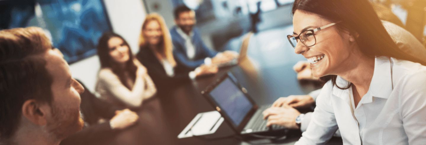 Groep collega's in overleg rondom een grote vergadertafel. Op de voorgrond zijn twee collega's geanimeerd in gesprek kijkend op een ipad