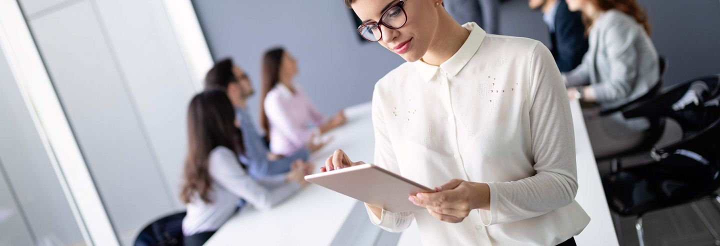 Medewerker die op Ipad staat te lezen. Op de achtergrond een groep collega's die aan vergadertafel in overleg zijn met elkaar.