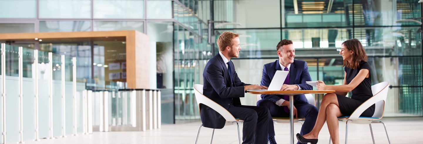 Drie collega's in een informeel overleg aan een tafel in een centrale ruimte van een kantoor.