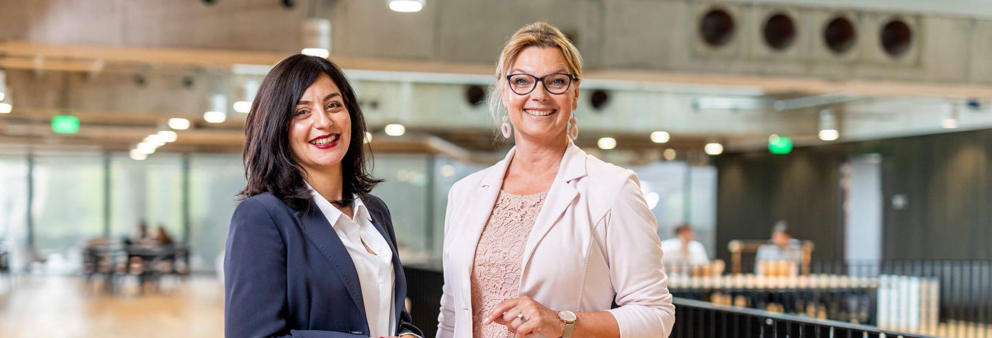 Twee vrouwen in zakelijke kleding staan geposeerd voor de camera in een lichte ruimte.