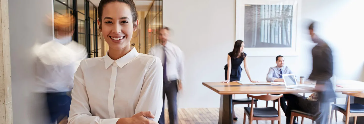 Vrouw staat middenin kantoorruimte waar verschillende collega's langslopen en aan het werk zijn.