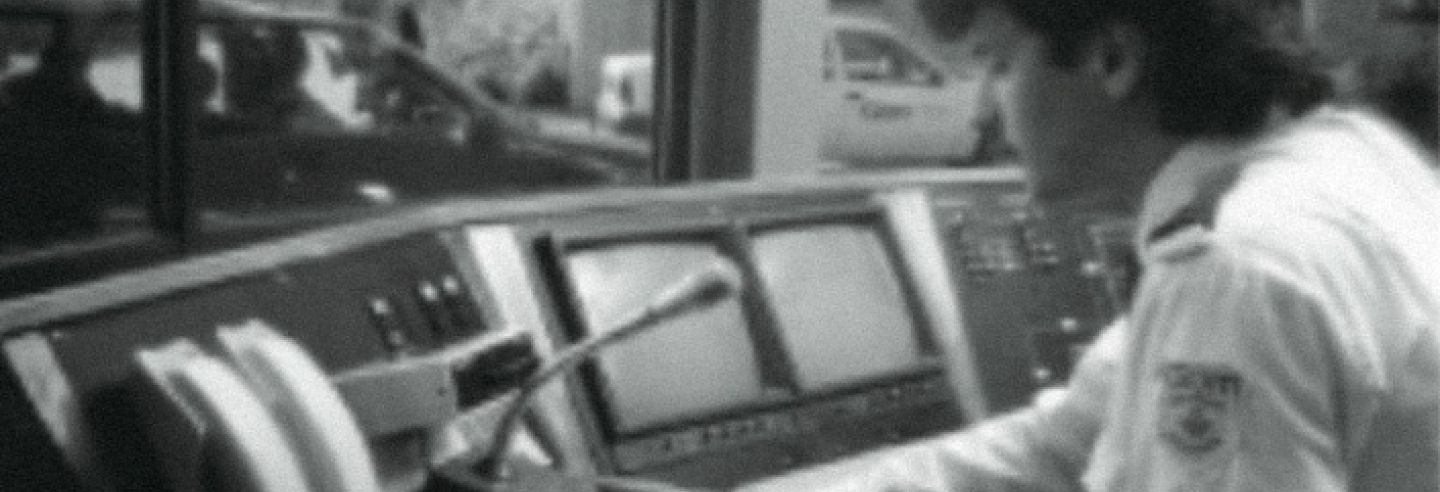 Zwart-wit foto met mannelijke beveiliger achter verouderde computerschermen sprekend in een microfoon. 