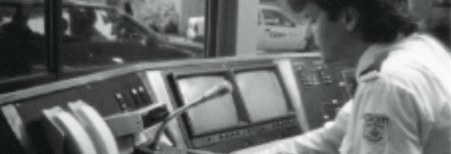 Zwart-wit foto met mannelijke beveiliger achter verouderde computerschermen sprekend in een microfoon. 