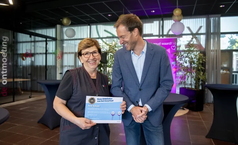 Schoonmaakster Ajsa heeft haar oorkonde vast, Jeroen Hofmeester staat naast haar en kijkt haar lachend aan