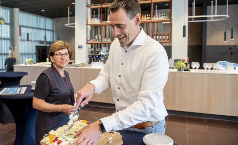 Collega snijdt de taart aan, terwijl schoonmaakster Ajsa voorbij loopt