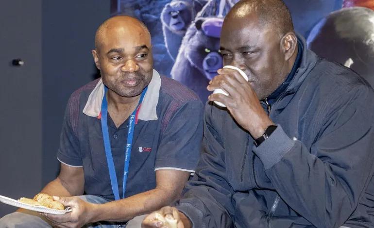 2 schoonmaakmedewerkers zitten naast elkaar en de een heeft een bordje met eten en de ander drinkt uit een bekertje