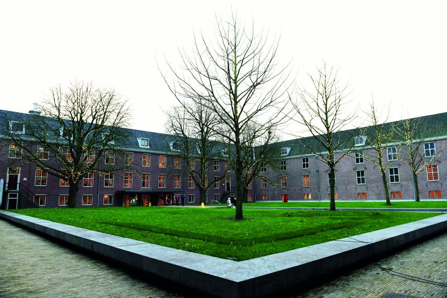 Dit is de binnentuin van de Hermitage in Amsterdam met een groot groen grasveld met verschillende grote bomen.