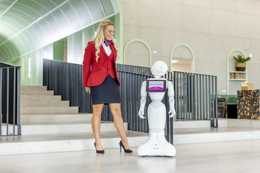 Gastvrouw  in uniform staat naar robot in centrale hal van een kantoorpand.