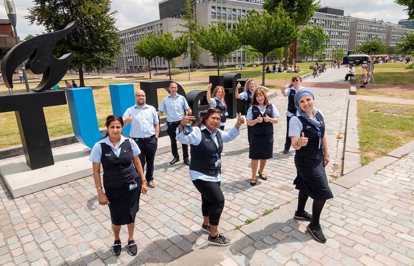 9 medewerkers van Gom Onderwijs staan voor de grote letters TU Delft op het campusterrein en steken hun duim omhoog