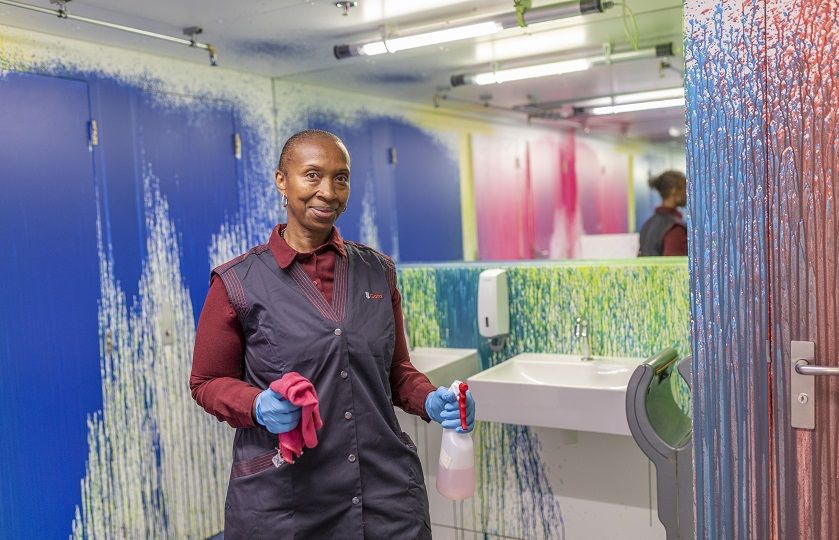 Maggy Lopes Robalo kijkt lachend in de camera en staat in het toilet wat is besproeid met verf door de watersprinklers