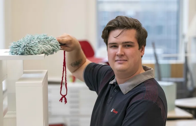 een medewerker van Gom Onderwijs kijkt in de camera en wist met een stofwisser de stof van een maquette