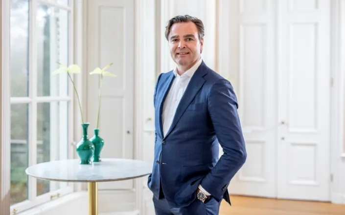 Zakelijk portret van een man in blauw pak die in een lichte kantoorruimte staat naast een tafel met twee groene vazen, passend bij een interview over de groei en successen van Gom in 2018.