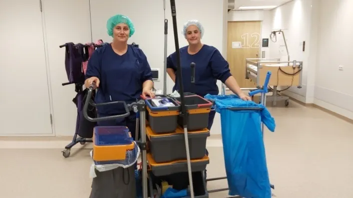 Claudia en Shea, schoonmaakmedewerkers bij Gom Zorg, staan met schoonmaakkar in de ziekenhuisgang klaar om de kamers te reinigen.