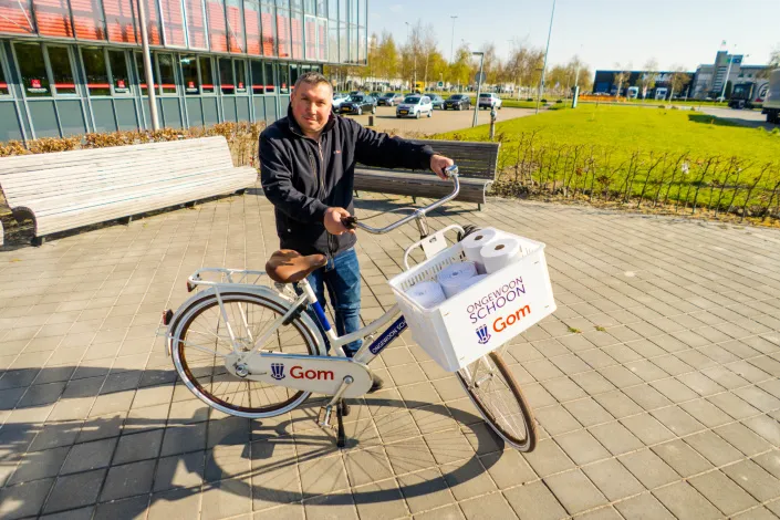 Medewerker met Gom-fiets op Schiphol, voorzien van mand met schoonmaakmaterialen, gebruikt voor efficiënte schoonmaak van bedrijfsgebouwen op de luchthaven.