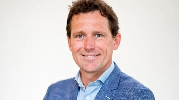 Portret van een man in een blauw colbert en lichtblauw overhemd, glimlachend tegen een neutrale achtergrond.