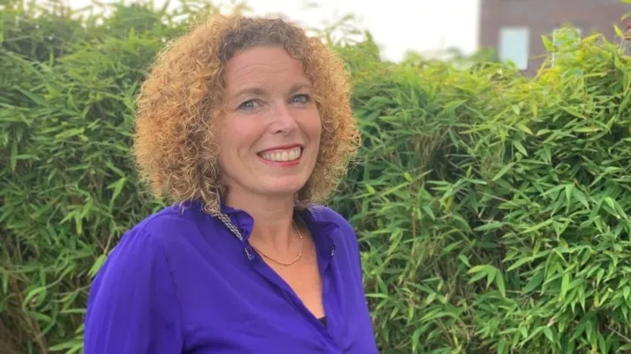 Portret van een vrouw met krullend haar en een paarse blouse, lachend voor een groene haag.