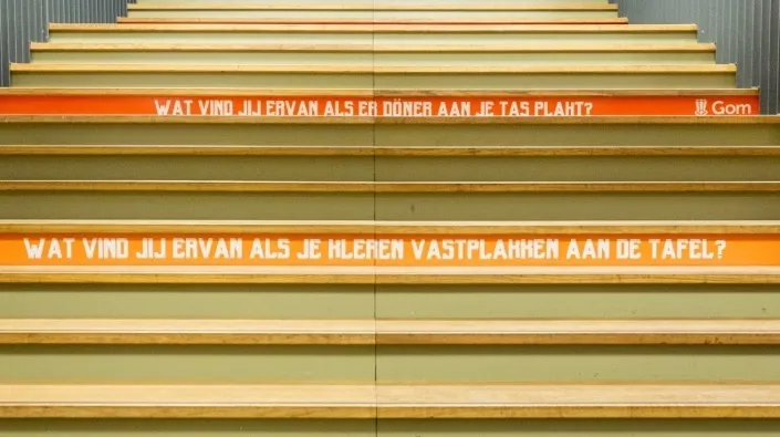 een brede houten trap in een school met op 2 achterborden teksen als 'Wat vind jij ervan als er döner aan je tas plakt?' en Wat vind jij ervan als je kleren aan de tafel vastplakken?'