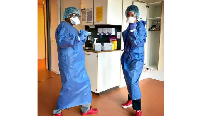 Twee zorgmedewerkers in blauwe beschermende kleding, mondmasker, handschoenen en haarnetje poseren in een ziekenhuiskamer.