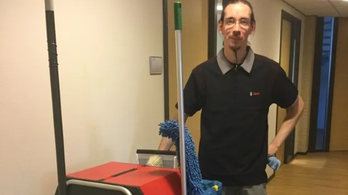Schoonmaker in Gom-uniform poseert met schoonmaakkar en dweil in een gang van een kantoorgebouw.