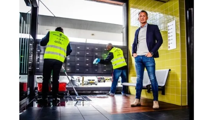 Twee schoonmakers in gele veiligheidshesjes maken een ruimte schoon, terwijl een man in pak en spijkerbroek toekijkt bij een muur met gele tegels.