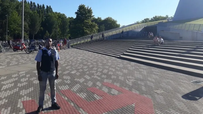 Beveiliger staat buiten op een plein met trappen bij een modern gebouw op een zonnige dag.