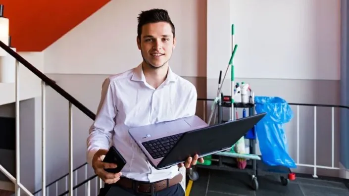 Mbo-student tijdens de facilitaire vakwedstrijd Skills Heroes met laptop en telefoon in een praktijkruimte met schoonmaakmaterialen op de achtergrond.
