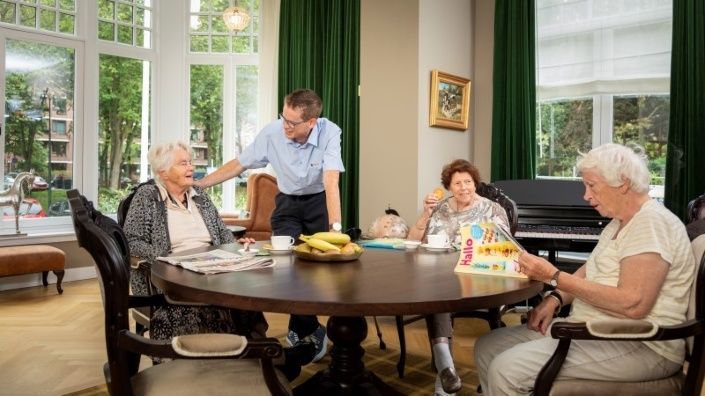 Schoonmaker van Gom houdt een praatje met drie ouderen die zitten aan een ronde tafel.