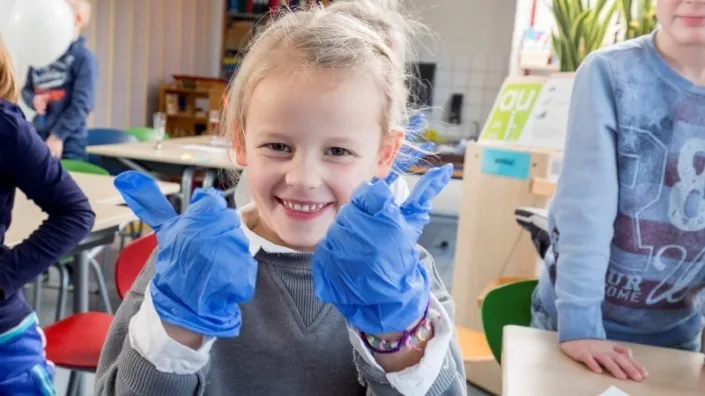 Kind in klaslokaal lacht en steekt duimen op terwijl het blauwe schoonmaakhandschoenen draagt, onderdeel van de proef met probiotica voor schonere schooltoiletten.