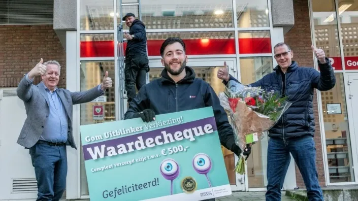Blije medewerker ontvangt bloemen en een grote waardecheque van €500 tijdens de Gom Uitblinker Verkiezing, omringd door collega’s die applaudisseren en duimen opsteken.