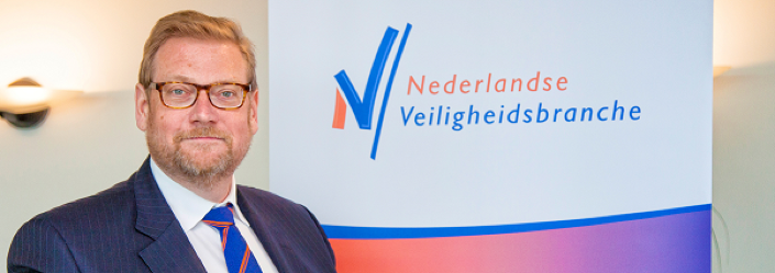 Man in pak staat voor een muur met daarop het logo van de Nederlandse veiligheidsbranche