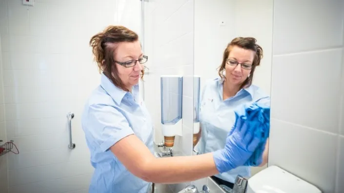 Schoonmaakmedewerker in lichtblauw uniform en handschoenen maakt een spiegel schoon in een sanitaire ruimte.