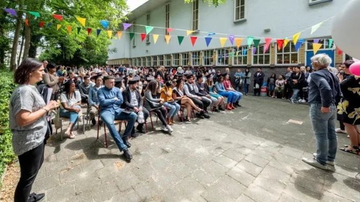 Groep studenten met afstudeerhoeden zit buiten tijdens een diploma-uitreiking, onder kleurrijke vlaggetjes, met publiek en begeleiders eromheen.