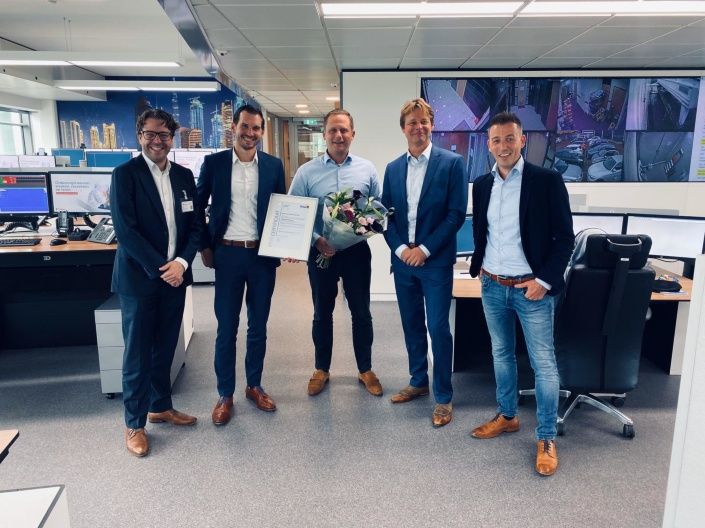 Vijf mannen in pak staan op de AlarmServiceCentrale geposeerd met een certificaat en bloemen in hun hand.