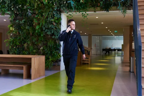 Mannelijke beveiliger loopt in zijn uniform door gang met een groene omgeving en belt met de centrale over de ingekomen melding via een portofoon.