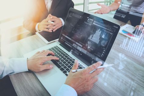 Twee mannen en een vrouw vergaderen aan tafel waarbij je meekijkt op de laptop van één van de mannen wie advies geeft aan de rest van het gezelschap.