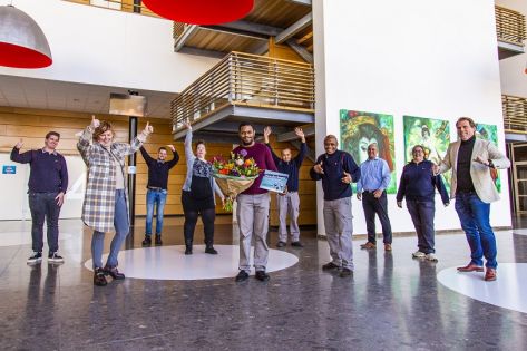 een groep van 8 collega's van Gom Offices staan juichend om Gom Uitblinker Edman Janga die een bos bloemen en een waardencheque in zijn handen heeft