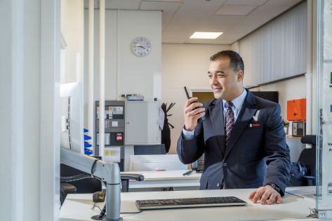 Mannelijke beveiliger in uniform praat via de portofoon met een collega beveiliger op locatie en kijkt daarnaast ook gelijk op zijn computerscherm achter zijn bureau.