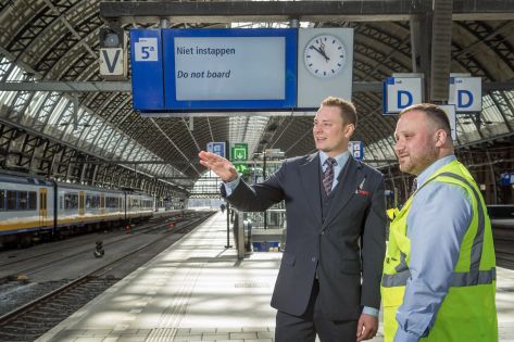 Mannelijke beveiliger in uniform legt aan mannelijke beveiliger met een veiligheidshesje uit wat de situatie is. Zij staan samen op het perron van een treinstation.