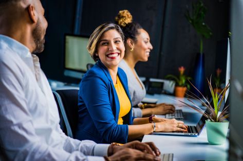 Een man en twee vrouwen zitten naast elkaar aan een bureau achter de computer, de focus ligt op een vrouw met een blauw jasje en een geel shirt.