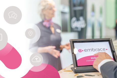 Je ziet de hand van een bezoeker die zich aanmeldt op een tablet met daarop het logo van Embrace. Achter de tablet zie je een Embrace-medewerkster in gesprek met een bezoeker.