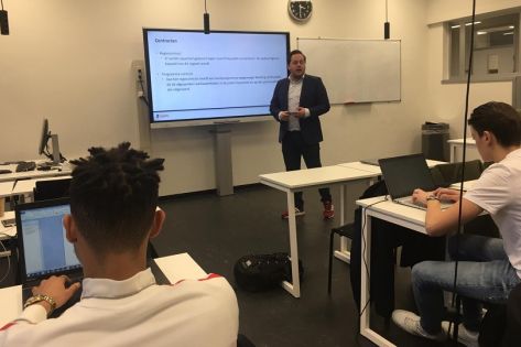 David van Boheemen staat als gastdocent voor een groot digitaal schoolbord, op de voorgrond zitten 2 studenten aantekeningen te maken