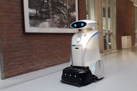 Schoonmaakrobot Ella, een witte robot met digitale ogen, rijdt in een gang voorbij een schilderij