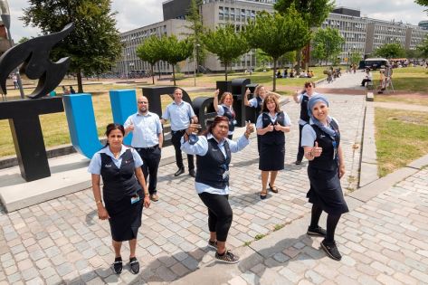 9 medewerkers van Gom Onderwijs staan voor de grote letters TU Delft op het campusterrein en steken hun duim omhoog