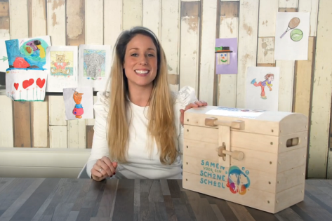 Marleen Lapidaire zit achter een tafel met daarop de houten leskist met het logo van Samen voor een Schone school erop