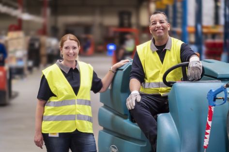 Twee schoonmaakmedewerkers van Gom Logistiek kijken lachend in de camera, 1 hiervan zit op een schrobzuigmachine, de ander staat ernaast