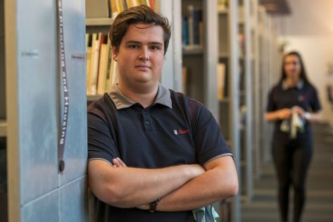 een medewerker van Gom Onderwijs leunt tegen een boekenkast en kijkt in de camera, op de achtergrond staat een vrouwelijke collega met een schoonmaakdoekje in haar hand