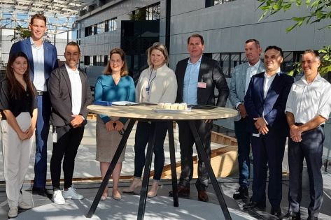 Een groep van 9 mensen staan om een tafel en kijken in de camera.